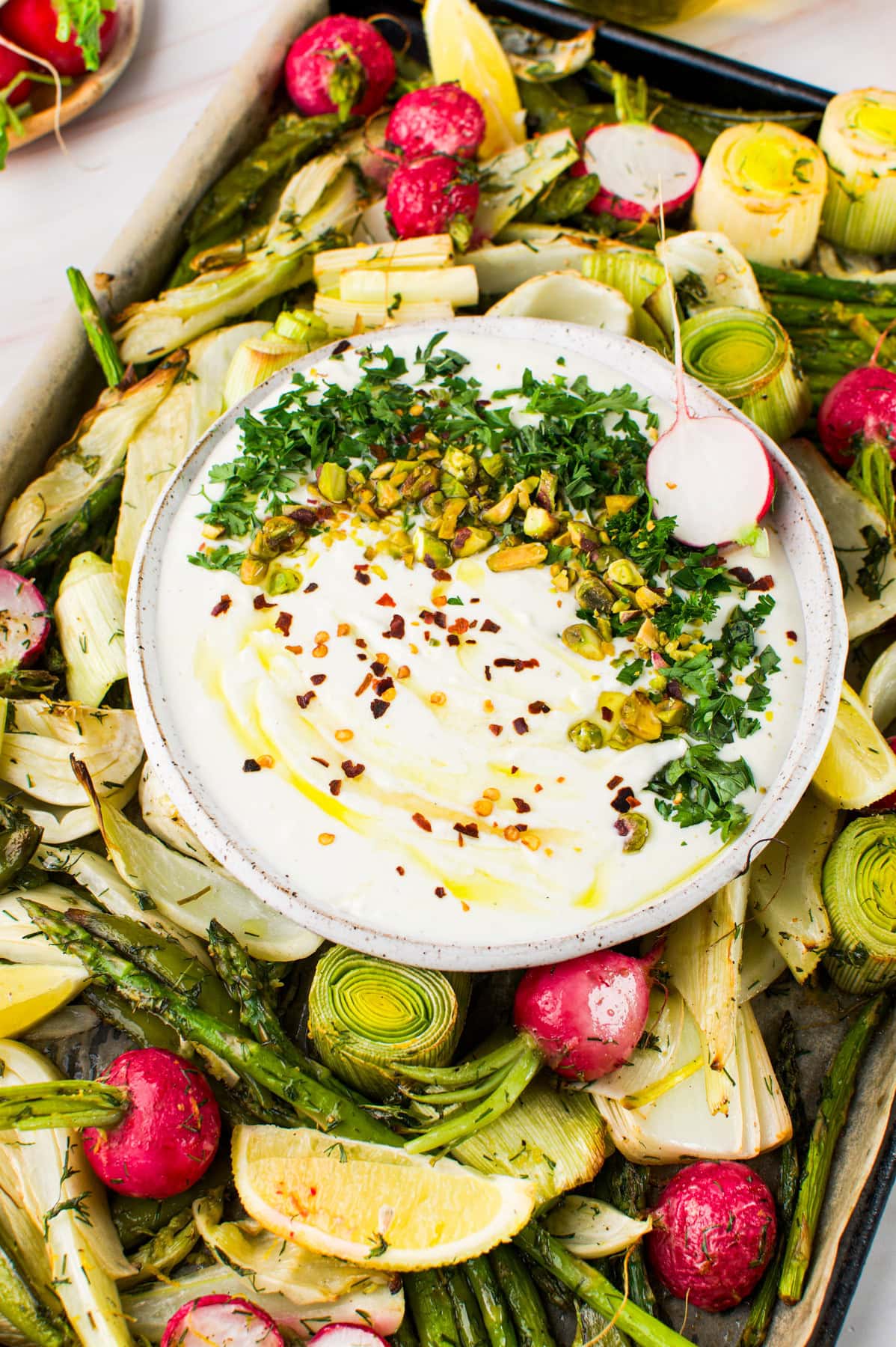A bowl of creamy dip topped with chopped pistachios, parsley, and red pepper flakes sits on a tray surrounded by roasted vegetables, including asparagus, radishes, leeks, artichokes, and lemon wedges.
