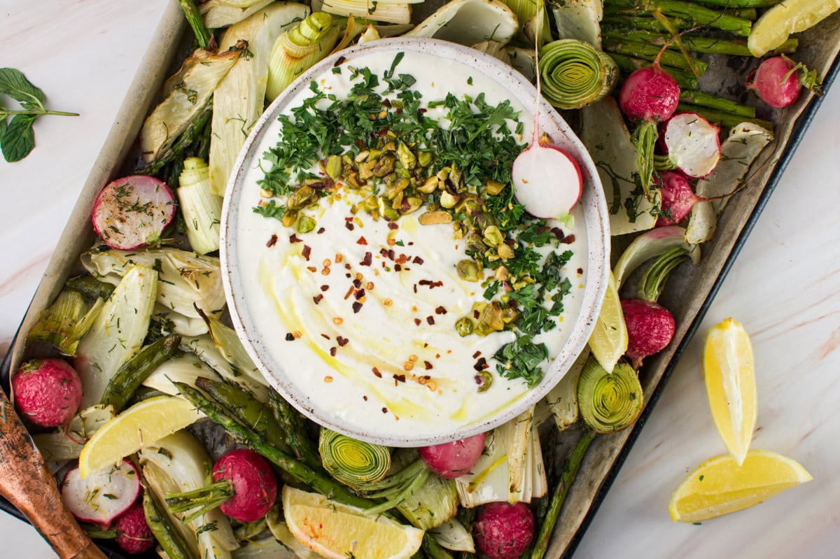 A bowl of creamy dip garnished with chopped herbs, nuts, and chili flakes sits on a tray with roasted vegetables, including radishes, leeks, asparagus, and fennel, with lemon wedges on the side.