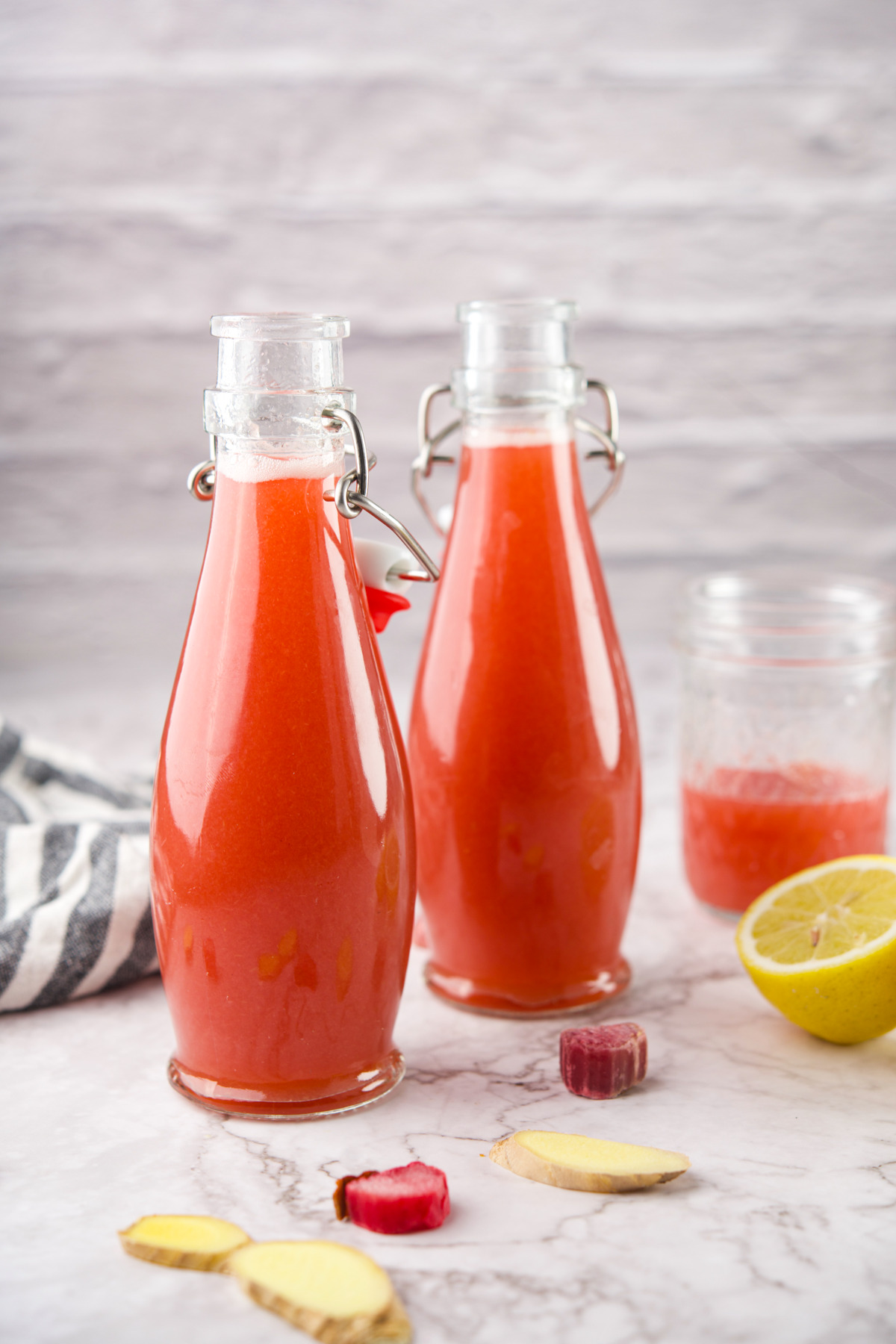 Two glass bottles filled with Homemade Rhubarb Syrup sit on a marble surface, next to a sliced lemon, ginger pieces, rhubarb, a striped towel, and a mason jar containing more juice. The background is textured and light-colored.