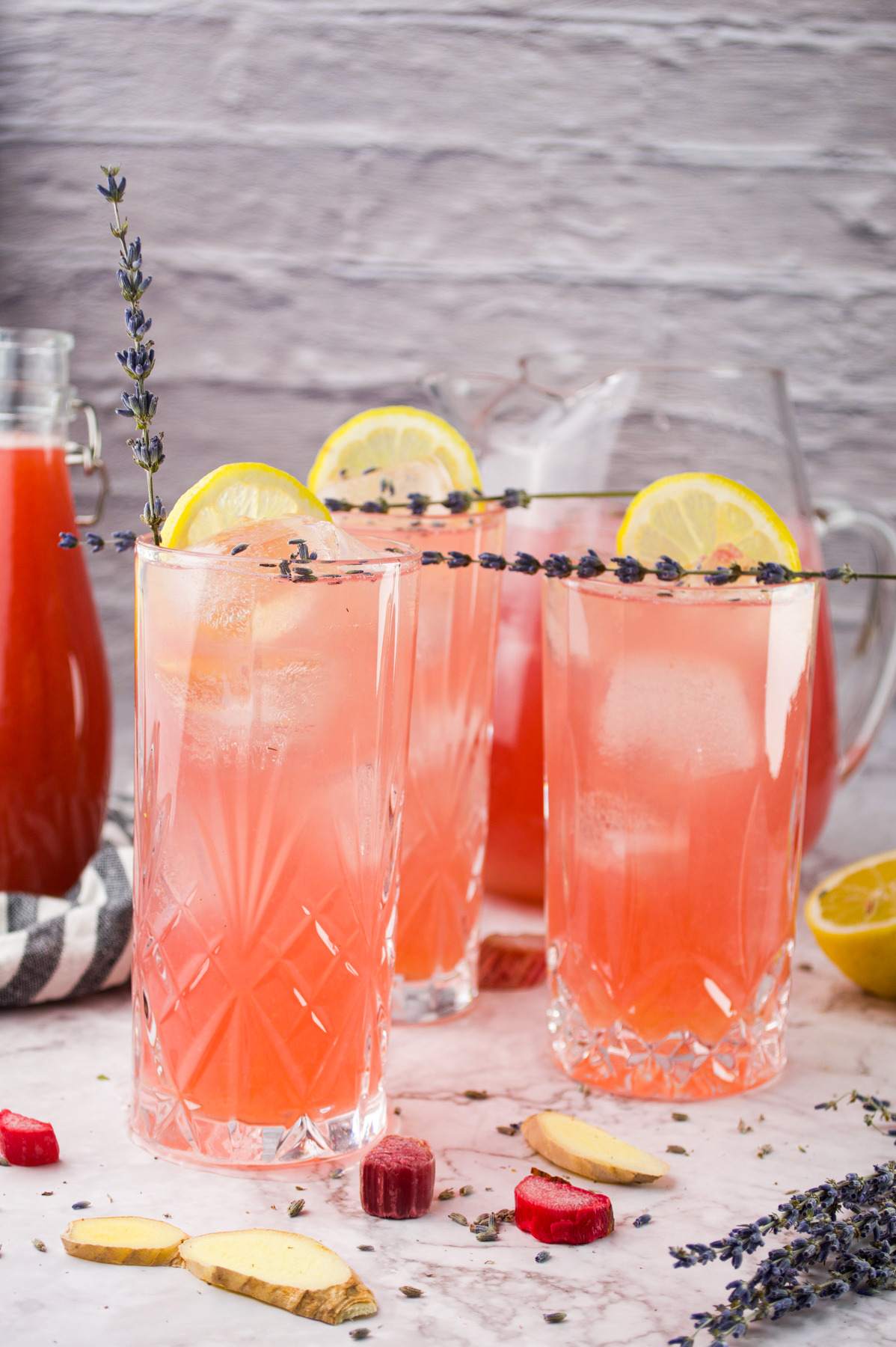 Three tall glasses of Rhubarb Lavender Lemonade with ice, garnished with lemon slices and sprigs of lavender, sit on a marble surface with fresh ginger, rhubarb slices, and a pitcher in the background.