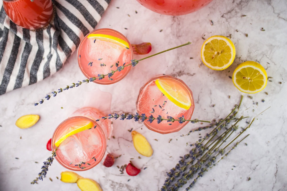 Three glasses of pink lemonade with ice, lemon slices, and lavender sprigs are on a marble surface, surrounded by lemon halves, ginger, strawberry slices, and lavender, next to a striped cloth.