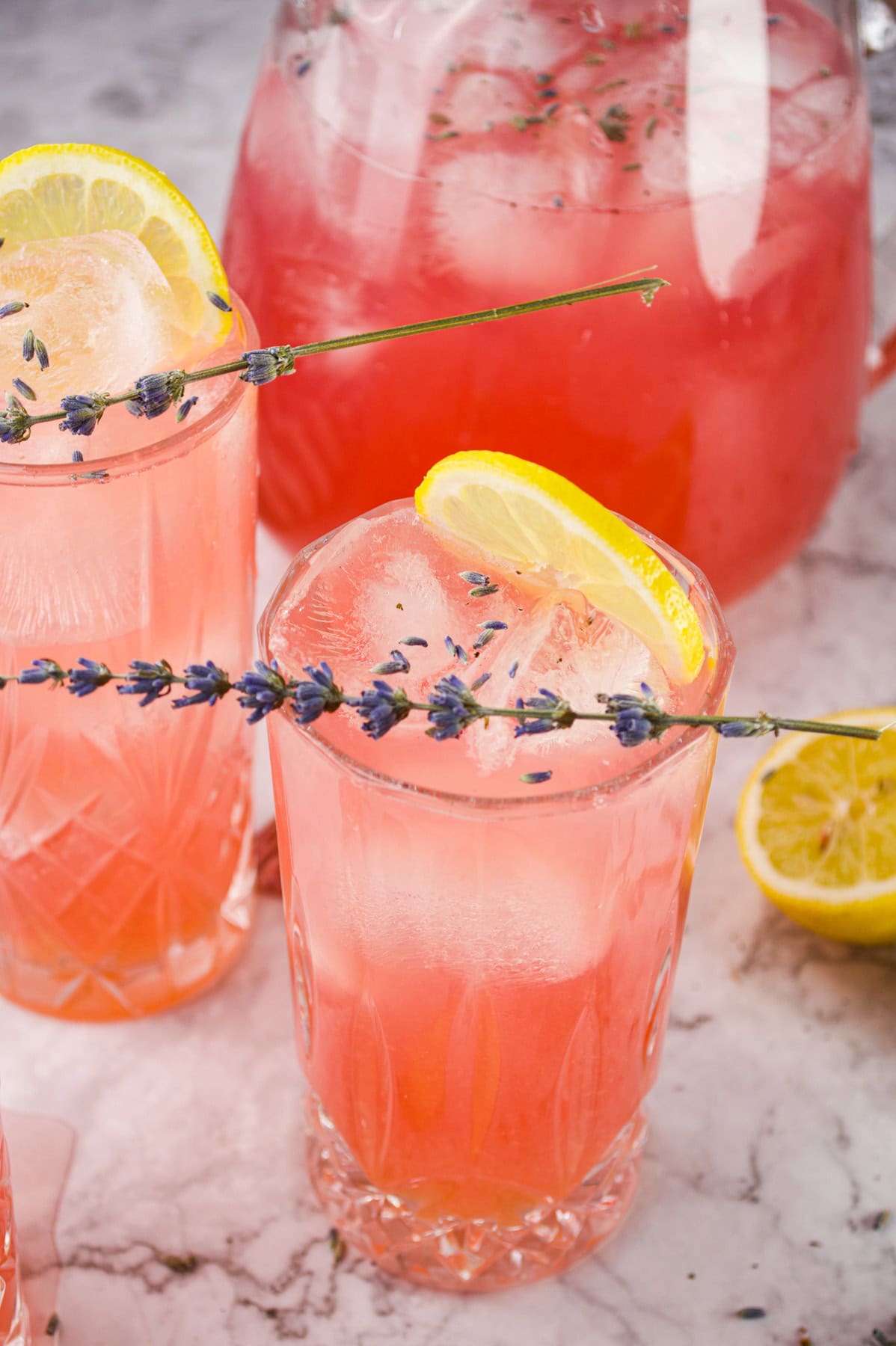 Two glasses of pink lemonade with ice, garnished with a lemon slice and a sprig of lavender, sit on a marble surface. A pitcher of the same drink and a halved lemon are in the background.