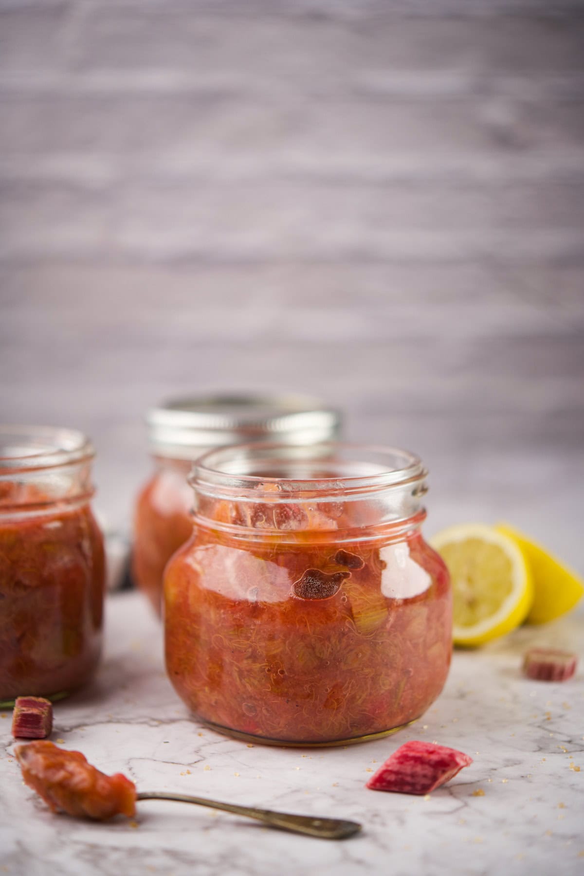 Three glass jars filled with homemade rhubarb compote sit on a light surface. One jar is open, and a halved lemon and scattered rhubarb pieces are nearby. The background is softly blurred.