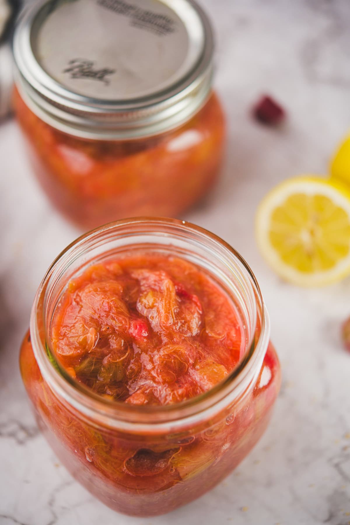 An open glass jar filled with chunky homemade fruit preserves, with another sealed jar and sliced lemons in the background on a light surface.