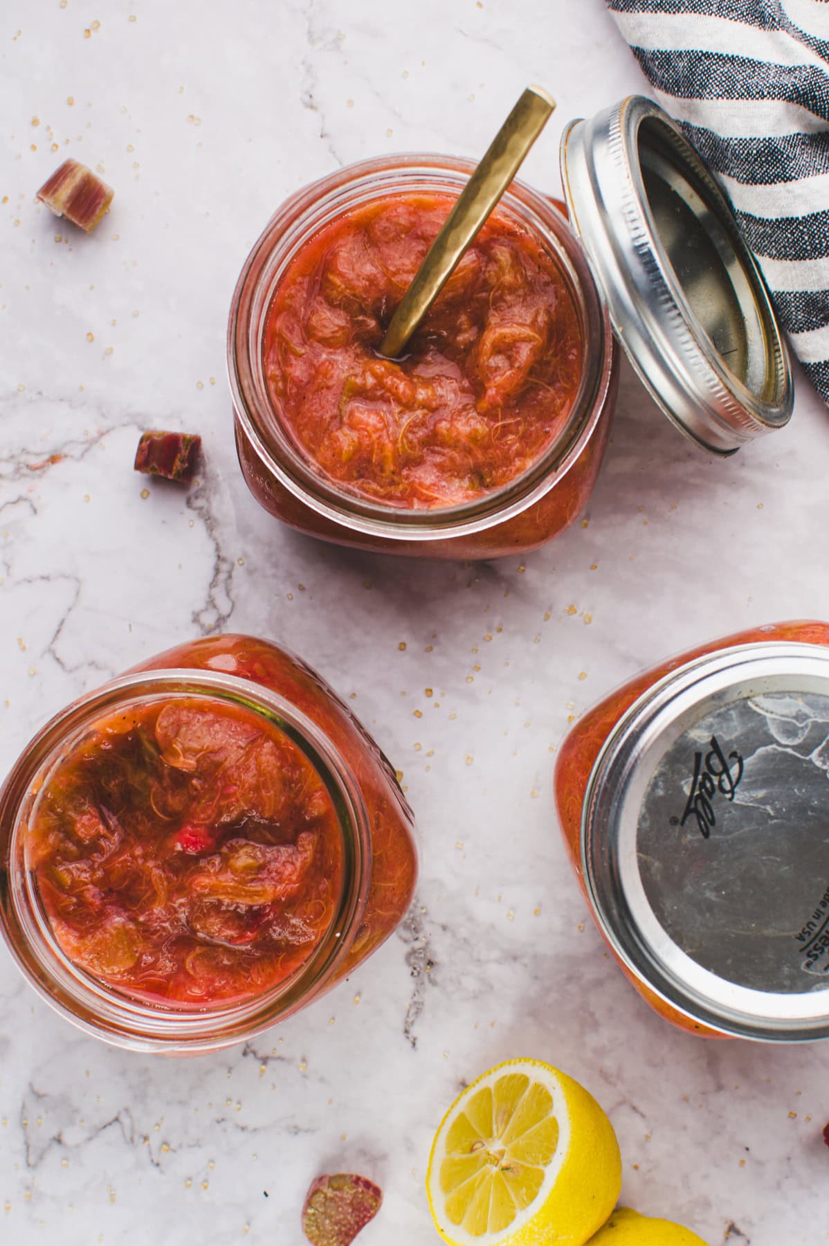 Overhead view of two jars filled with chunky rhubarb preserves, one with a spoon. Two jar lids, lemon halves, and pieces of rhubarb are nearby on a marble surface with a striped cloth in the corner.