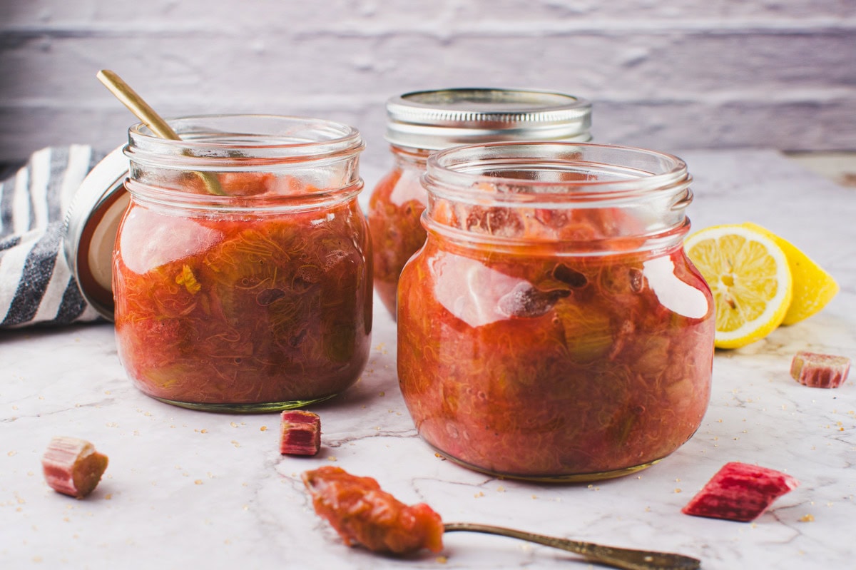 Three glass jars filled with homemade rhubarb jam sit on a marble surface. One jar is open with a spoon inside, while the others are closed. Sliced rhubarb and a lemon wedge are nearby. A striped cloth is in the background.