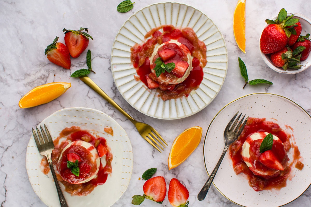 Three plates of panna cotta with strawberry sauce, fresh mint, and strawberries are arranged on a marble surface with orange slices, more strawberries, and a small bowl of strawberries surrounding them. Forks rest on two plates.