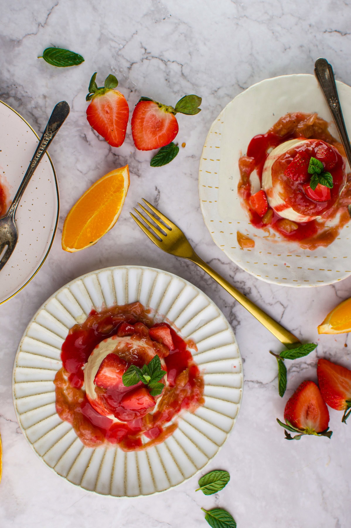 Three plates with vanilla ice cream topped with strawberry sauce and fresh mint, surrounded by sliced strawberries, orange wedges, and mint leaves on a marble surface. A gold fork lies between the plates.