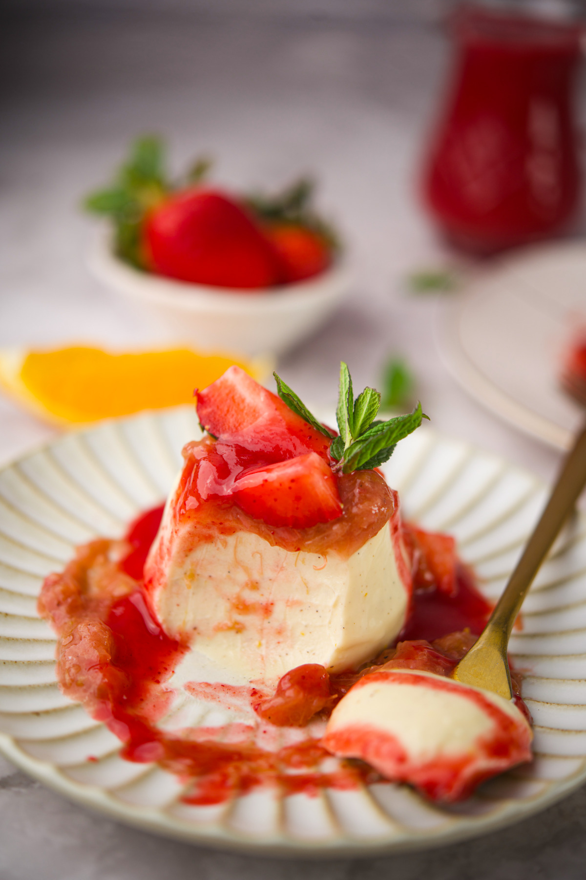 A Pannacotta With Rhubarb Compote strawberry sauce and fresh strawberries, garnished with a mint sprig on a striped plate. A gold spoon rests beside it. In the background are strawberries and a jar of sauce.