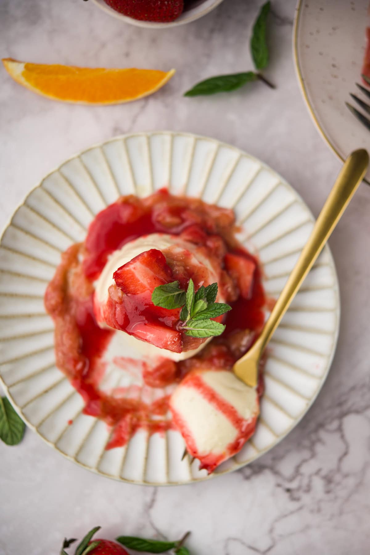 A plate with vanilla ice cream topped with strawberry and rhubarb sauce, garnished with fresh mint leaves. A gold spoon rests on the side. Orange slice, strawberries, and mint leaves are scattered nearby.