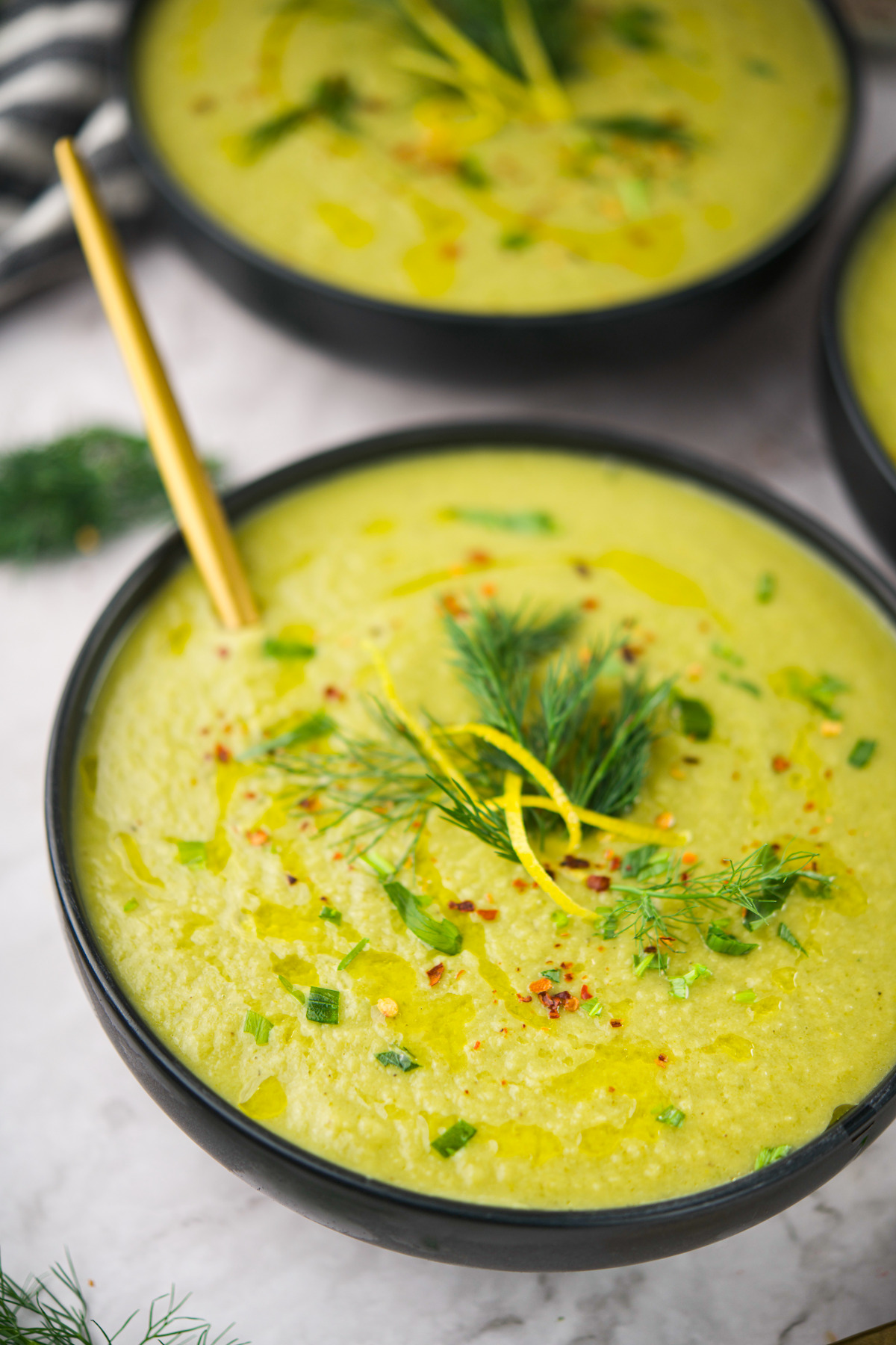 A black bowl filled with creamy green soup, garnished with fresh dill, chopped herbs, lemon zest, and a drizzle of olive oil. A gold spoon rests inside the bowl. Another bowl is partially visible in the background.