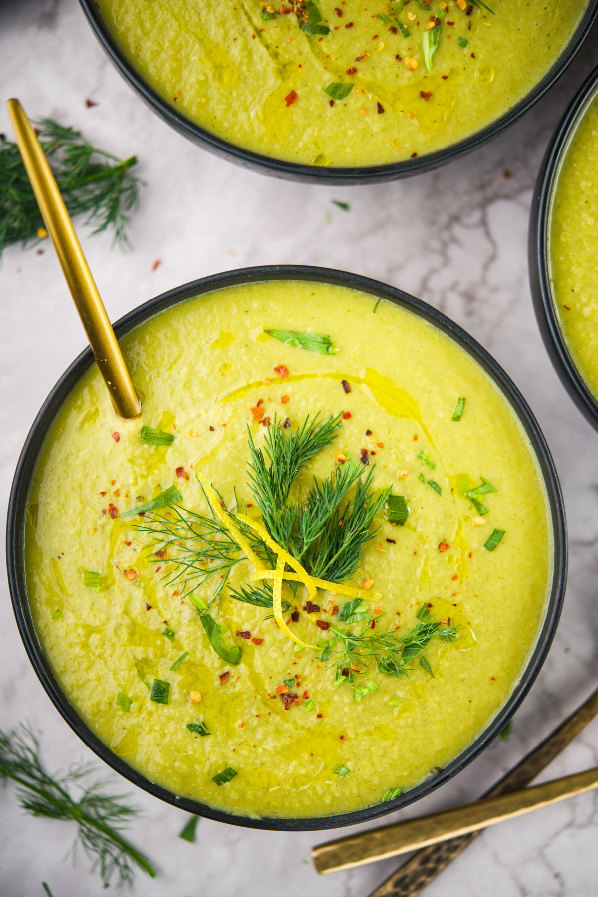 A bowl of Fennel Leek Soup garnished with fresh dill, chopped herbs, chili flakes, lemon zest, and a drizzle of olive oil, with a gold spoon resting inside the bowl.
