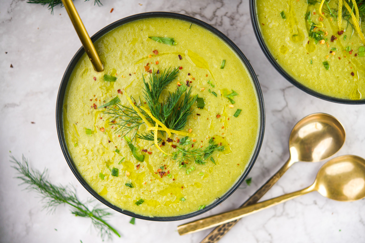 A bowl of creamy green soup garnished with fresh dill, lemon zest, and herbs, with a gold spoon resting inside. Two more gold spoons and sprigs of dill are placed nearby on a light marble surface.