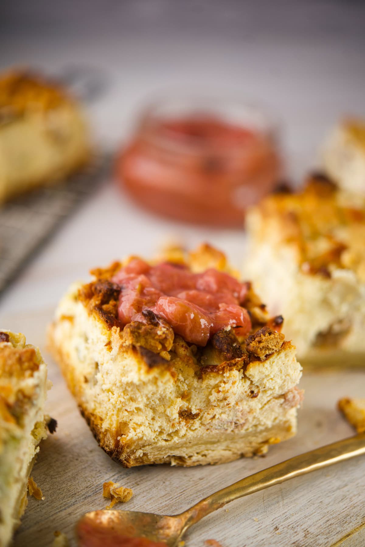 A close-up of a Rhubarb Cheesecake Bars placed on a light surface next to a gold fork. A blurred jar of compote and more cake slices are in the background.