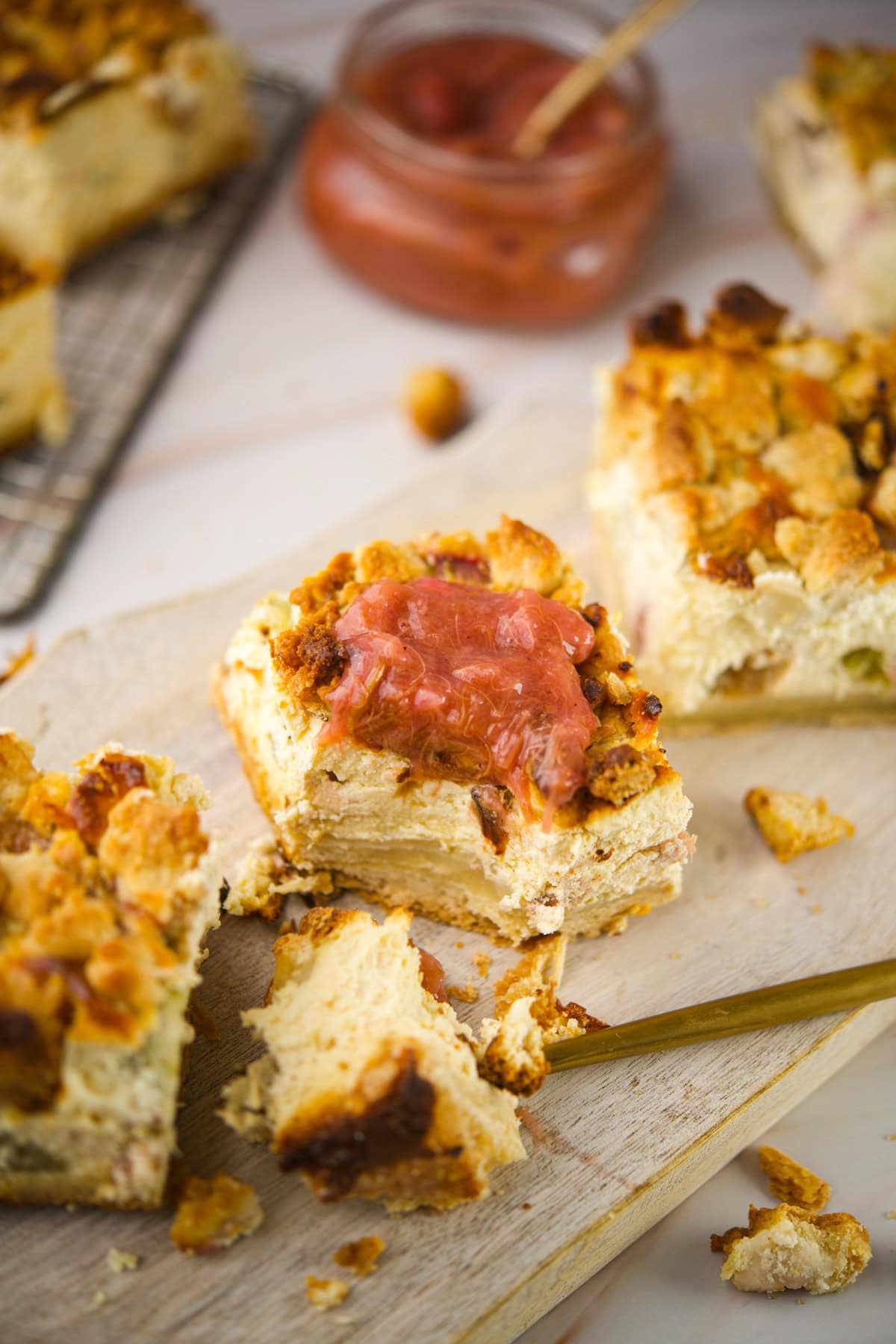 A close-up of crumbly rhubarb cheesecake bars on a wooden board, with one bar topped with pink fruit compote. More bars and a jar of compote are in the background. Crumbs are scattered around.
