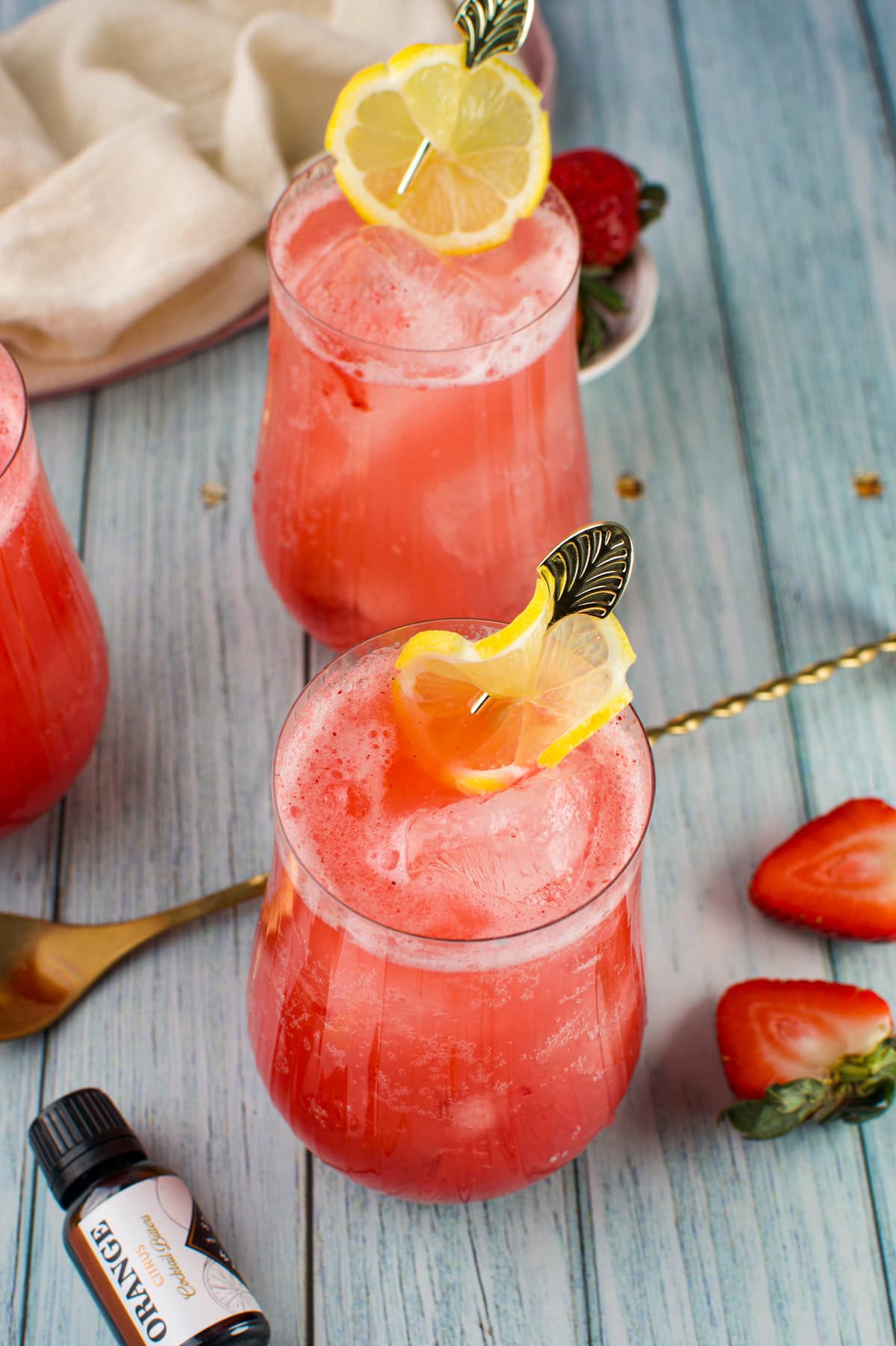 Three glasses of pink strawberry lemonade with ice are garnished with lemon slices. Fresh strawberries and a bottle labeled orange are on a blue wooden table beside the drinks.