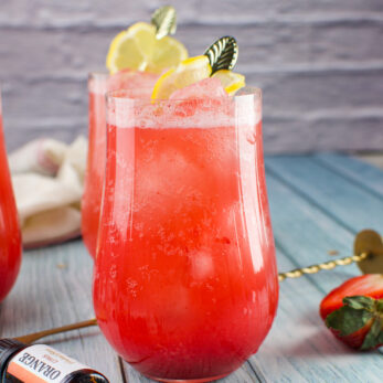 A vibrant Rhubarb Strawberry Spritzer in a clear glass, garnished with lemon slices and a decorative pick, sits on a blue wooden surface. Nearby are a strawberry, a cocktail stirrer, and a small bottle labeled orange.