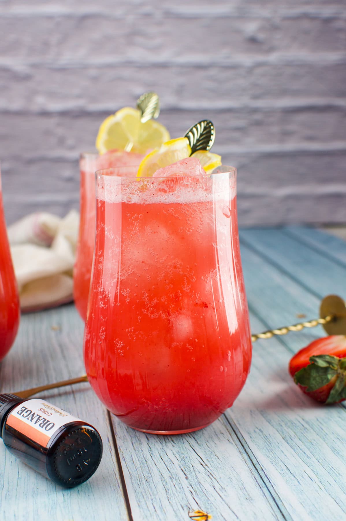 A vibrant Rhubarb Strawberry Spritzer in a clear glass, garnished with lemon slices and a decorative pick, sits on a blue wooden surface. Nearby are a strawberry, a cocktail stirrer, and a small bottle labeled orange.