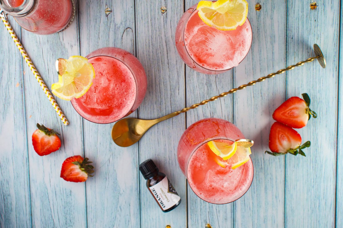 Three glasses of pink strawberry drinks with ice and lemon slices sit on a blue wooden surface, surrounded by fresh strawberries, a gold cocktail spoon, a striped straw, and a small flavoring bottle.