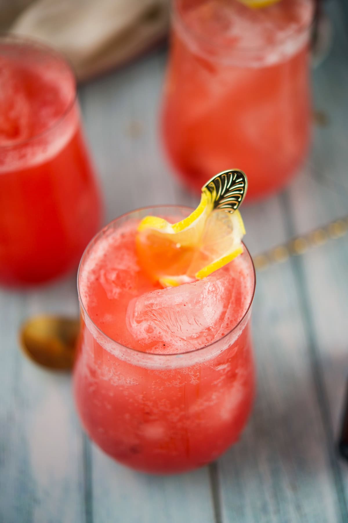 A close-up of a pink iced drink garnished with a lemon slice on the rim, served in a stemless glass on a light wooden surface; other similar drinks are blurred in the background.