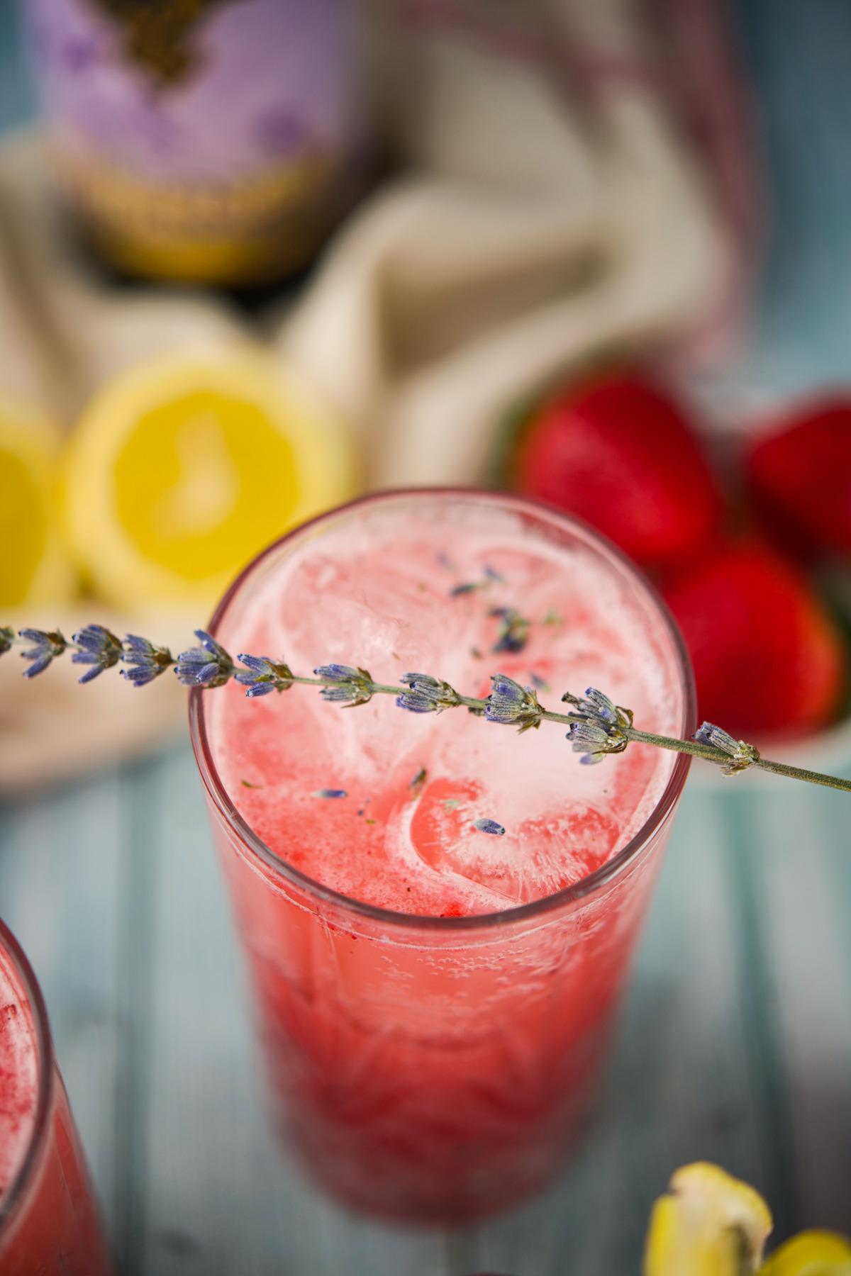 A close-up of a Strawberry Lavender Spritzer Cocktail , garnished with a sprig of lavender. Fresh strawberries and halved lemons are blurred in the background.