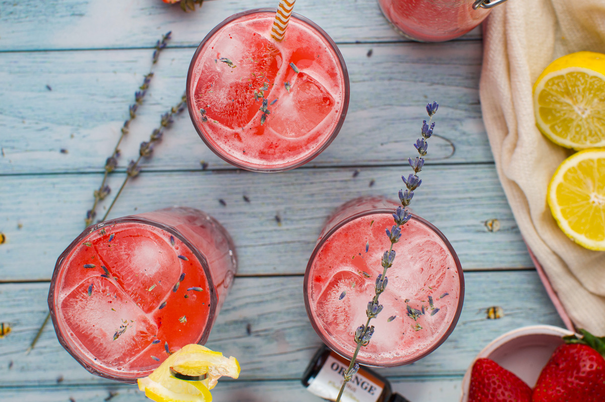 Three glasses of Strawberry Lavender Spritzer Cocktail with lavender sprigs and lemon garnishes on a blue wooden table, surrounded by sliced lemons, strawberries, and a napkin.