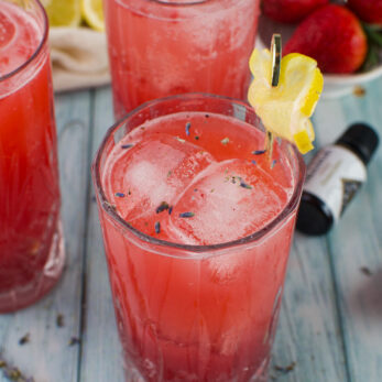 Three glasses of pink Strawberry Lavender Spritzer Cocktail with ice are garnished with lemon slices and lavender sprigs, set on a light blue wooden surface with fresh strawberries, lemon slices, and lavender nearby.