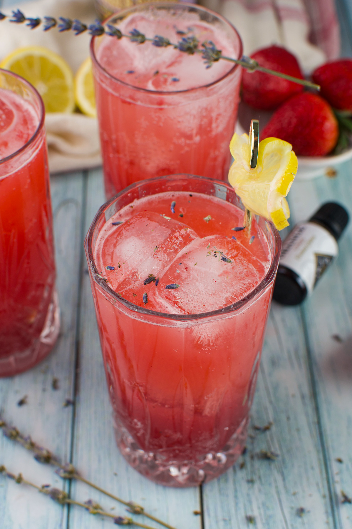 Three glasses of pink Strawberry Lavender Spritzer Cocktail with ice are garnished with lemon slices and lavender sprigs, set on a light blue wooden surface with fresh strawberries, lemon slices, and lavender nearby.