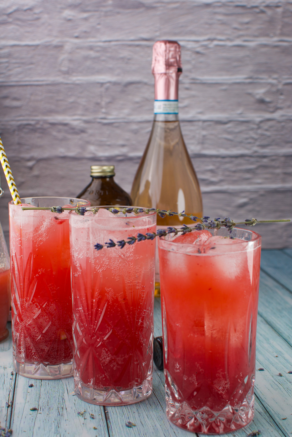 Three Strawberry Lavender Spritzer Cocktails with ice and lavender sprigs are served in tall crystal glasses on a blue wooden surface, with a bottle of sparkling wine and a brown bottle in the background. A gold-striped straw sits in one glass.