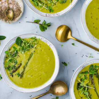 Four bowls of Roasted Asparagus, Leek, Cannellini Bean Soup garnished with herbs and asparagus, placed on a light surface with gold spoons nearby. A small dish with salt and pepper sits in the corner, surrounded by fresh mint leaves.