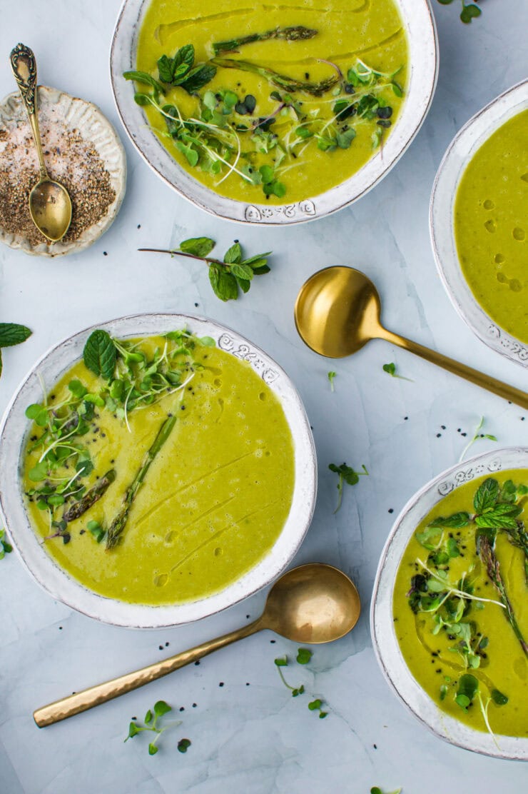 Four bowls of Roasted Asparagus, Leek, Cannellini Bean Soup garnished with herbs and asparagus, placed on a light surface with gold spoons nearby. A small dish with salt and pepper sits in the corner, surrounded by fresh mint leaves.