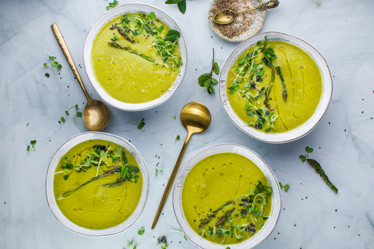 Four bowls of green soup, garnished with asparagus tips and fresh herbs, are arranged on a light marble surface with two gold spoons and scattered microgreens.