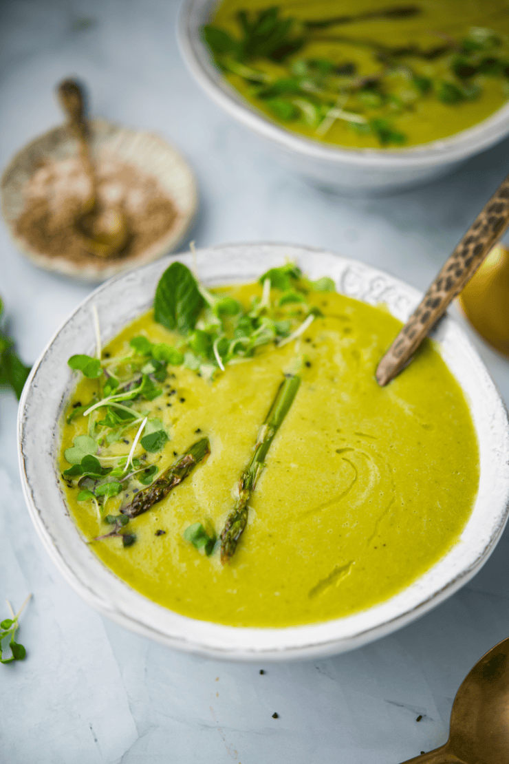 A bowl of creamy green soup garnished with asparagus spears, microgreens, and a mint leaf, with a spoon in the bowl. Another bowl and a dish of seasoning are visible in the background.