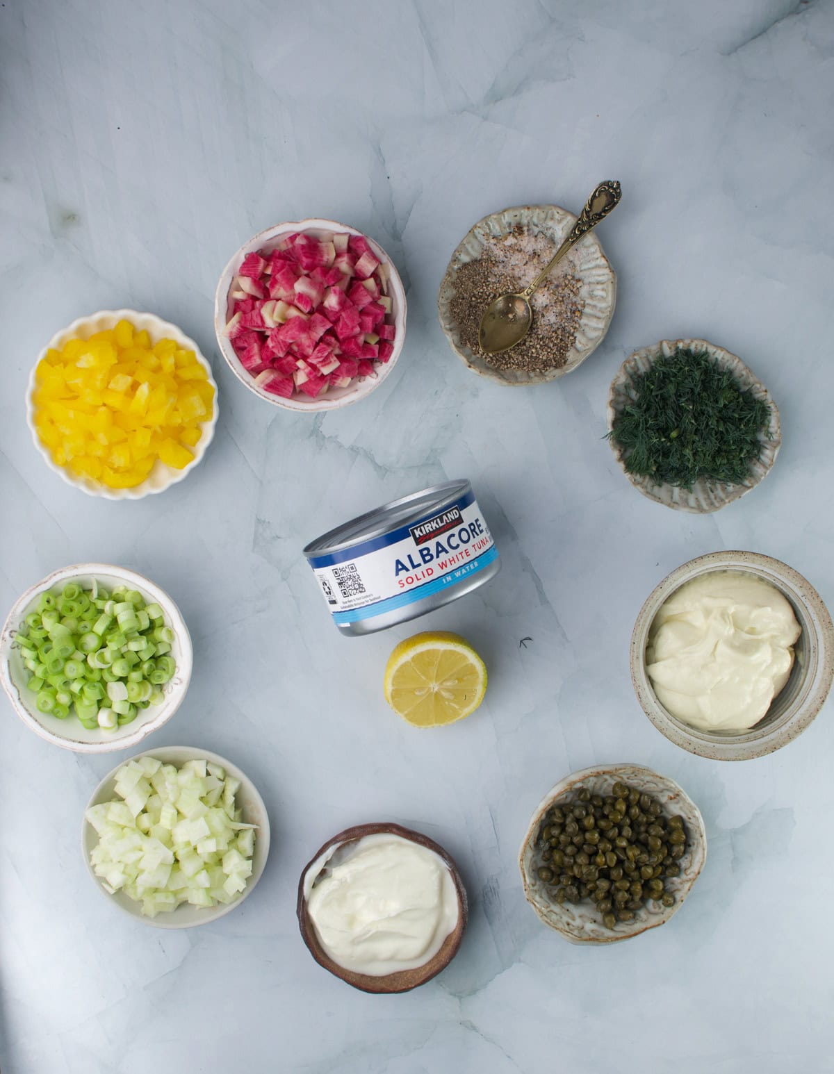 A can of albacore tuna surrounded by bowls of diced yellow bell pepper, radishes, green onion, cucumber, capers, fresh dill, seasoning, mayonnaise, and half a lemon on a marble surface.