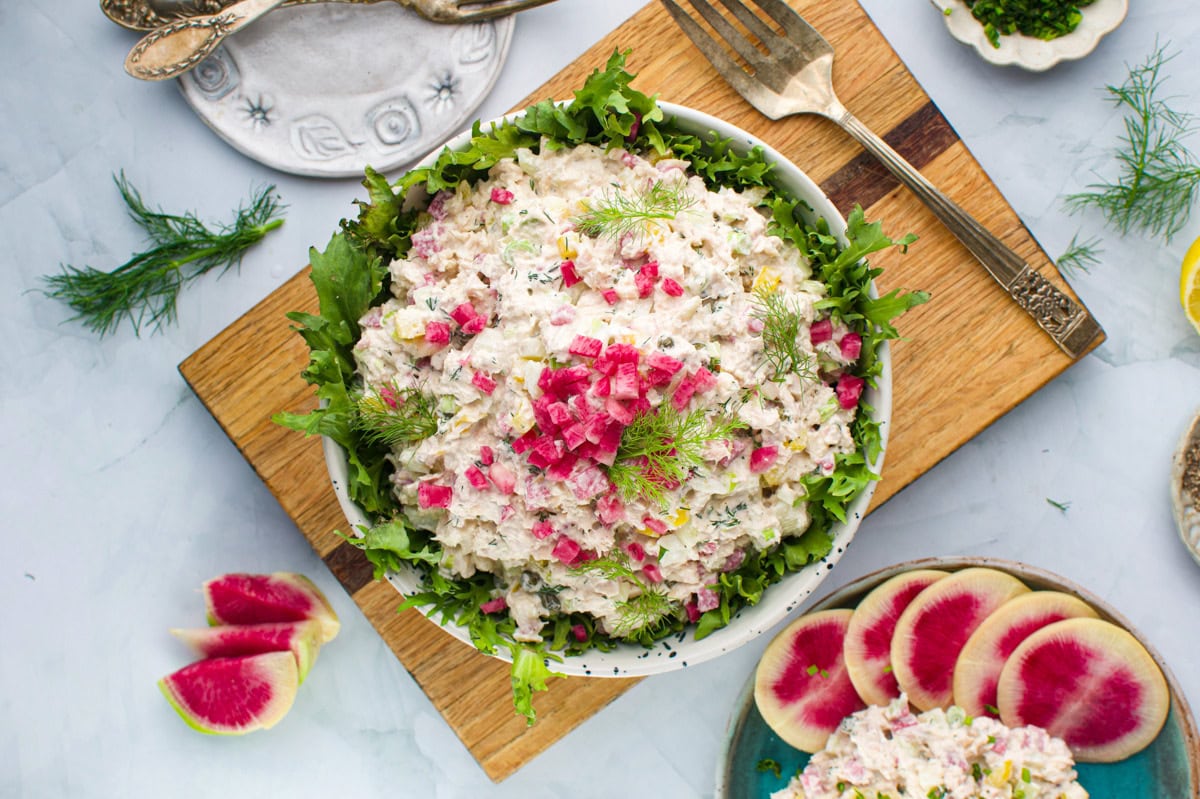 A bowl of creamy salad with chopped red onions and dill, served on leafy greens. Slices of radish and herbs surround the bowl on a wooden board, with plates, a fork, and a wedge of lemon nearby.