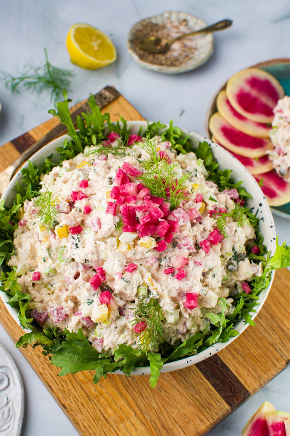 A bowl of creamy Spring Tuna Salad sits on a bed of leafy greens, garnished with pink radish cubes and fresh dill, on a wooden board. Slices of watermelon radish, a lemon half, and herbs surround the bowl.