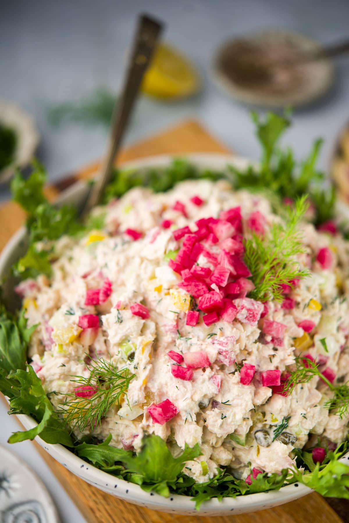 A bowl of creamy salad garnished with bright pink diced vegetables and fresh dill, surrounded by leafy greens, with a serving spoon and blurred background elements.