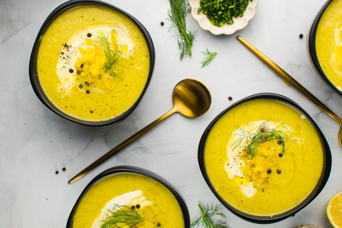 Four bowls of creamy yellow soup topped with herbs, cream, black pepper, and a drizzle of oil, sit on a white surface with gold spoons and garnish dishes nearby. Fresh herbs and a lemon slice are also visible.