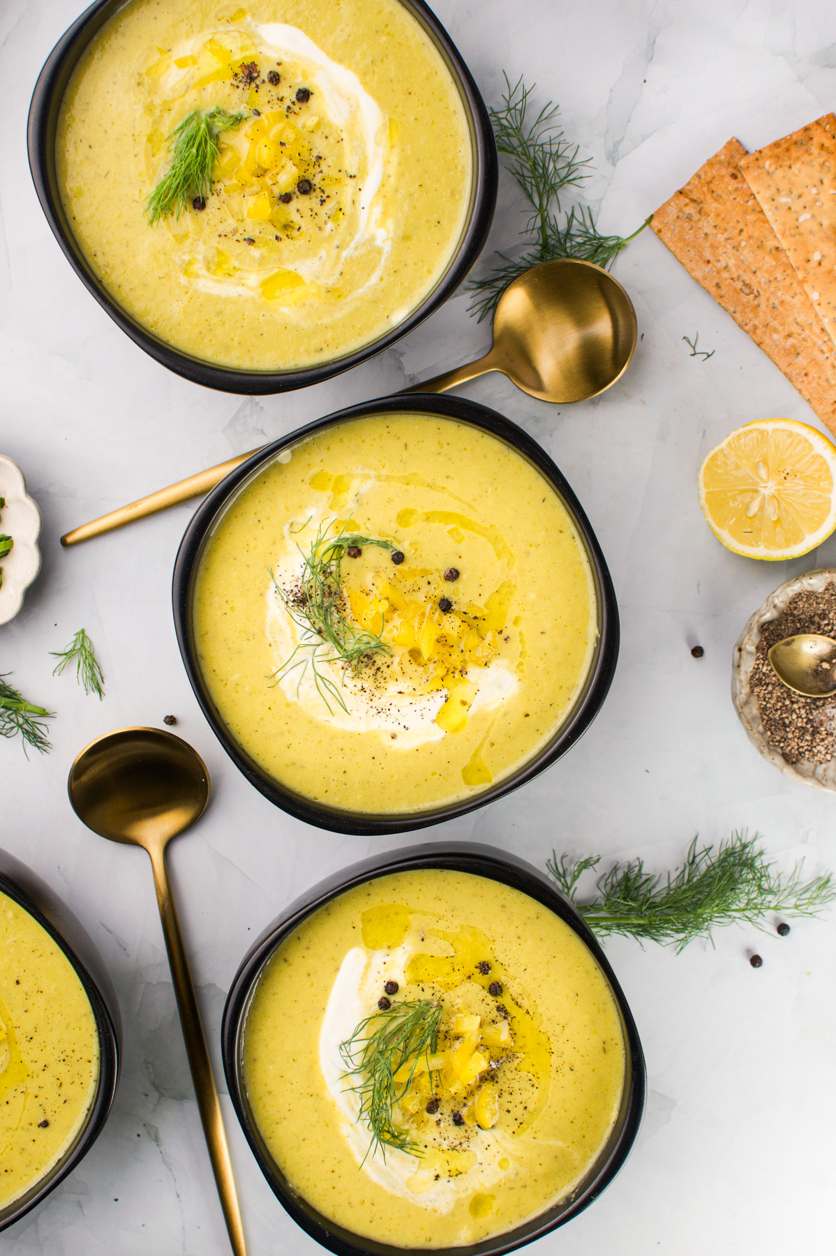 Three bowls of creamy yellow soup garnished with cream, olive oil, pepper, and fresh dill, are arranged on a marble surface with gold spoons, crisp crackers, a lemon half, dill sprigs, and spices nearby.