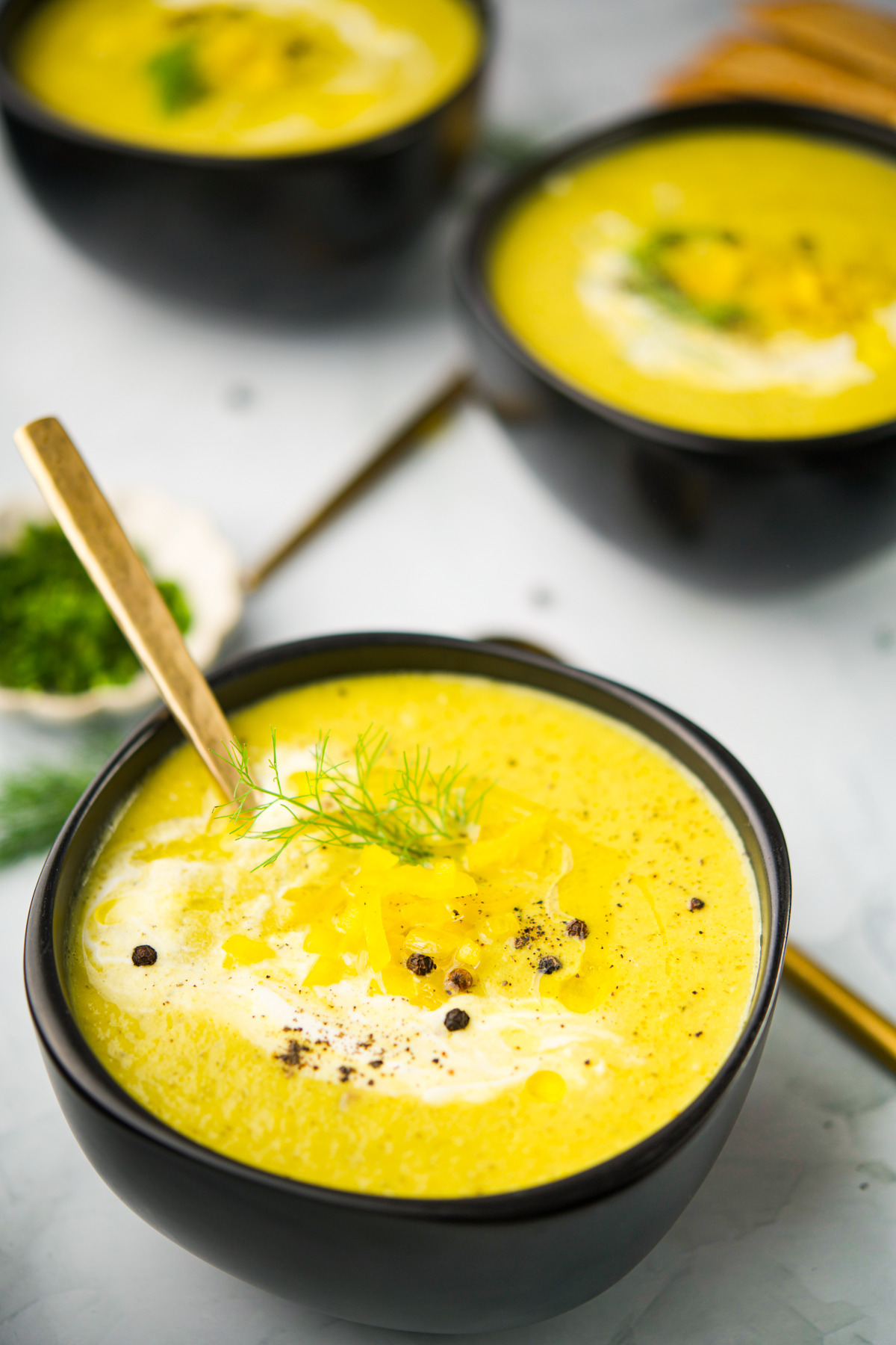 A close-up of a black bowl filled with Cream Of Fennel Pepper Dill Soup garnished with cream, black pepper, herbs, and a drizzle of oil. Two similar bowls of soup and a spoon are visible in the background.