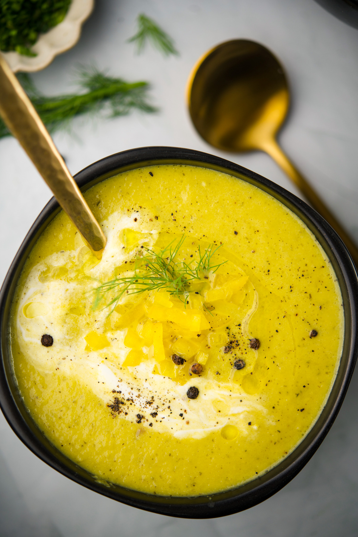 A bowl of creamy yellow soup garnished with fresh dill, diced yellow vegetables, black pepper, and a swirl of cream, with a gold spoon resting inside. A gold spoon and herbs are visible in the background.