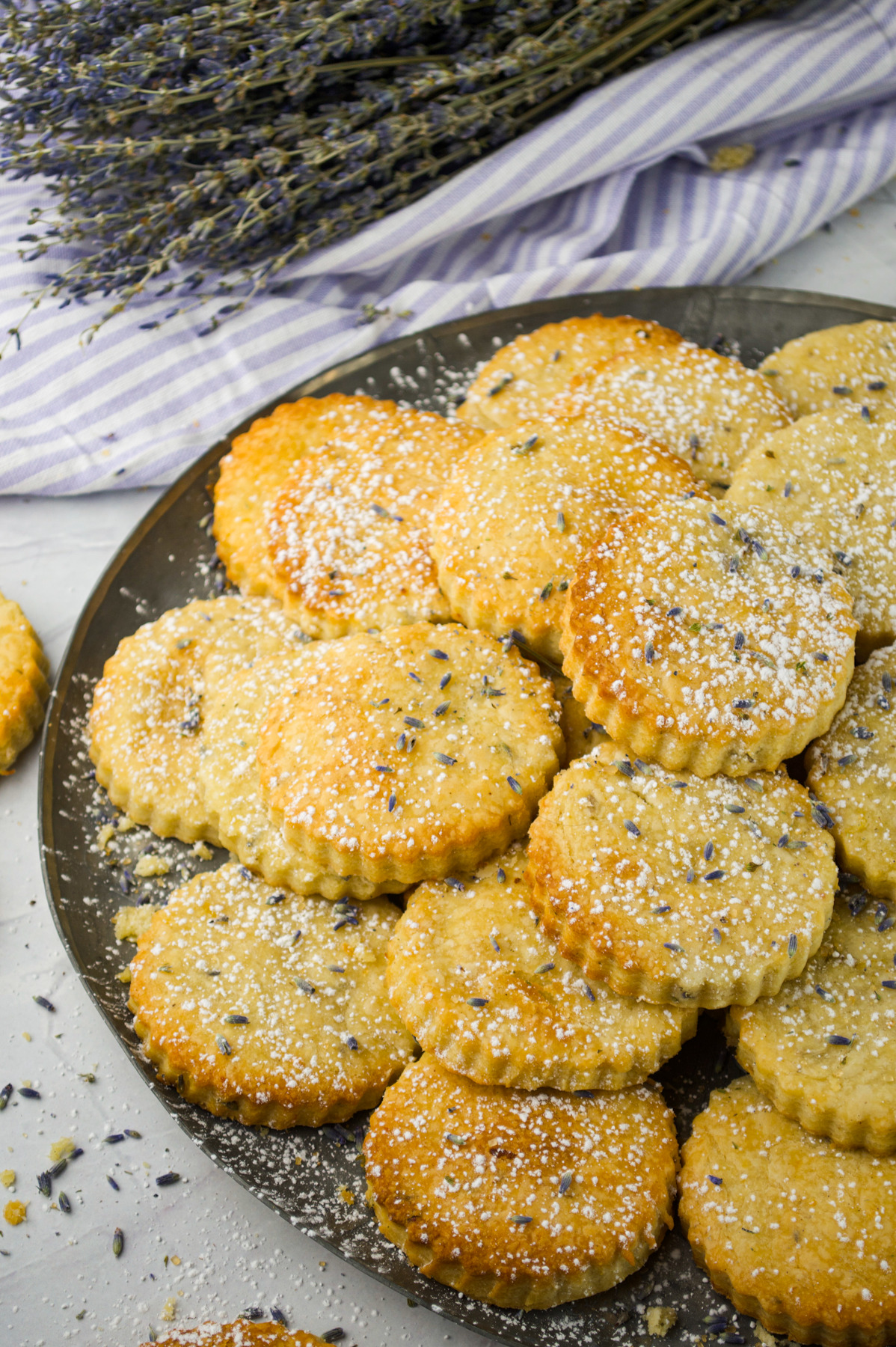 A plate of round lavender cookies sprinkled with powdered sugar sits on a light surface, with sprigs of dried lavender and a striped cloth in the background.
