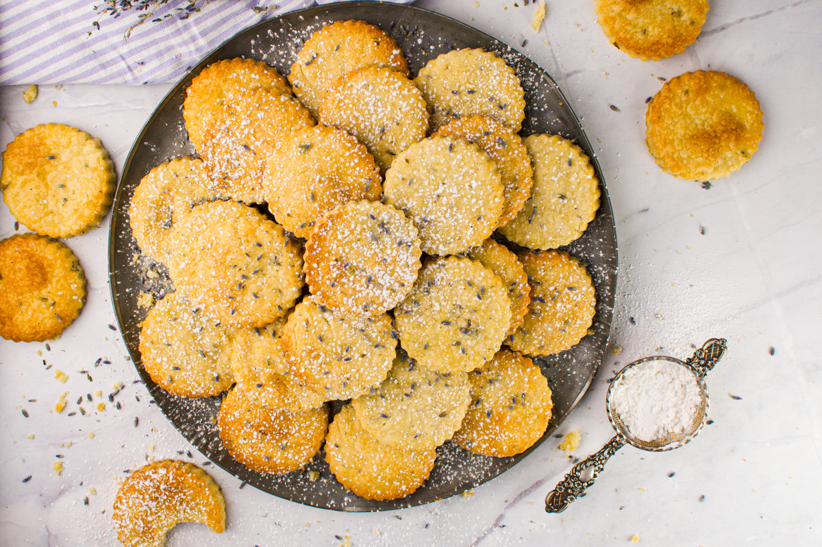 A plate piled with round, golden cookies sprinkled with powdered sugar sits on a marble surface. Some cookies and a small dish of powdered sugar are scattered around the plate. Lavender buds are also visible.