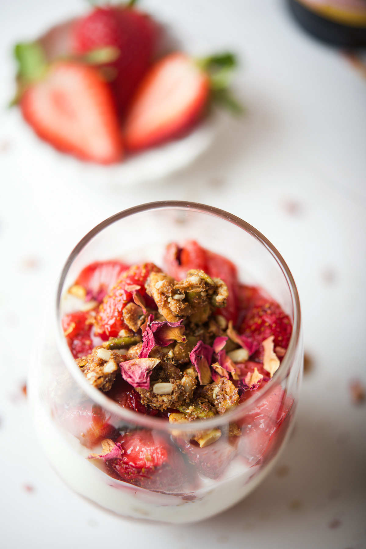 A glass filled with Rhubarb Strawberry Parfait with fresh strawberries, granola, and sprinkled with rose petals. In the background, sliced strawberries are visible on a small white plate.