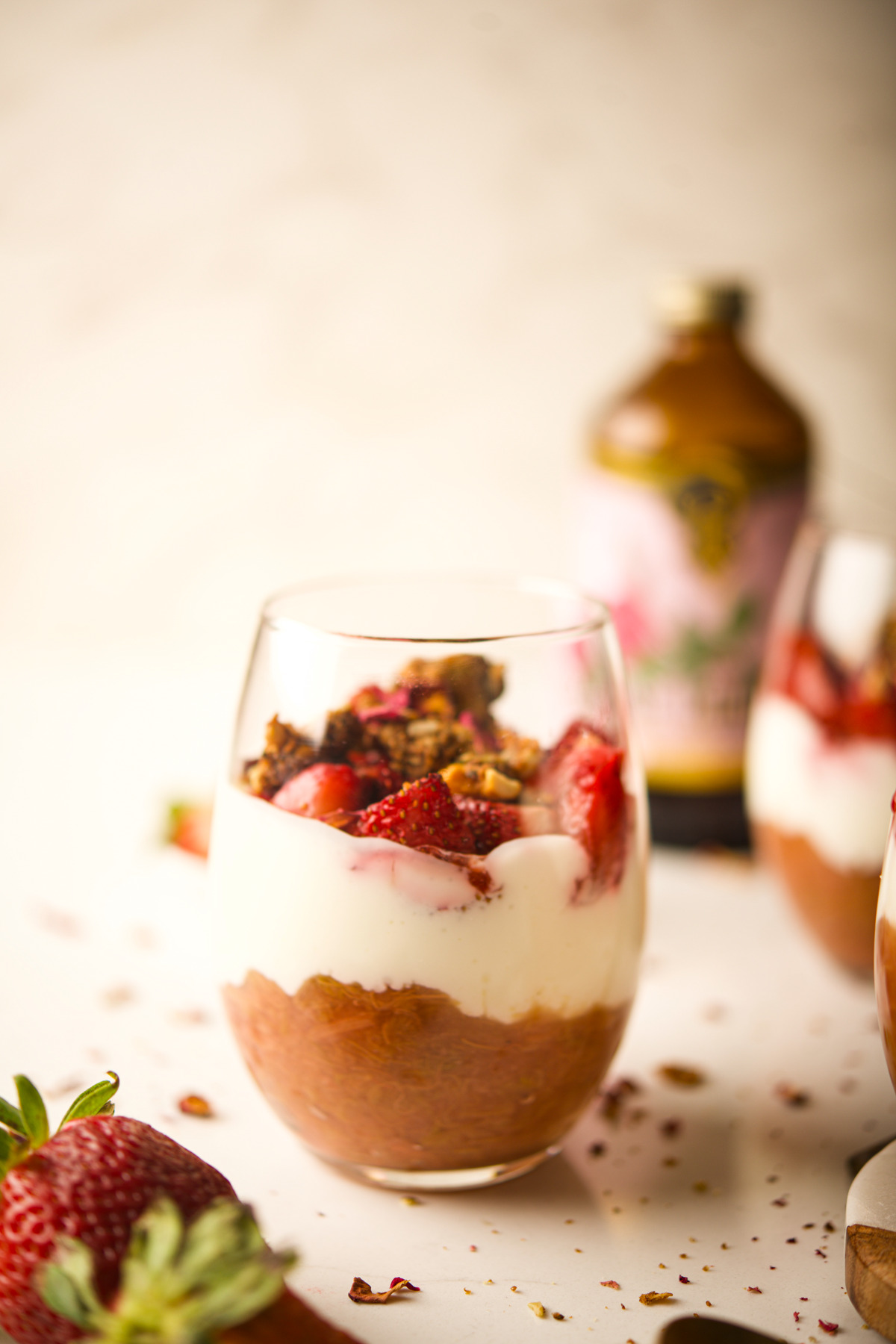 A glass filled with layers of Rhubarb Strawberry Parfait, creamy yogurt, granola, and fresh strawberry slices, with a blurred bottle and another glass in the background. A whole strawberry sits in the foreground.