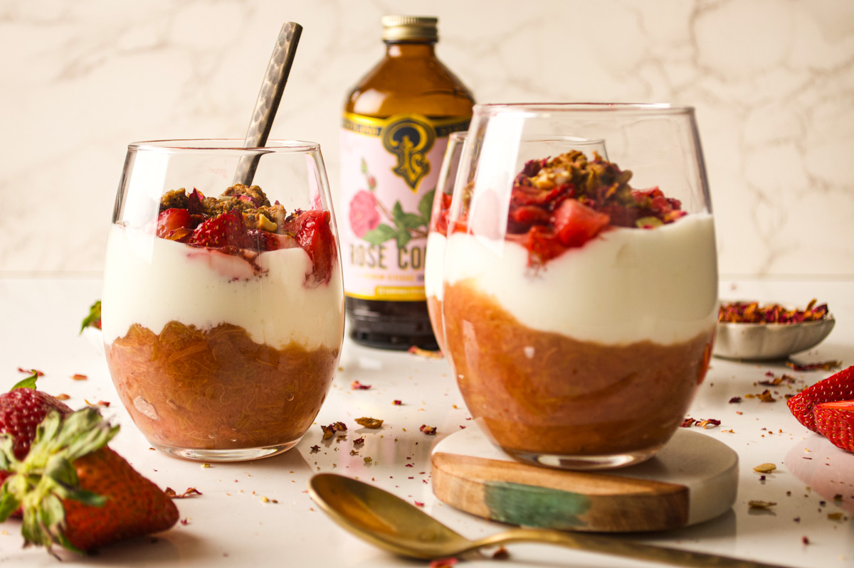 Two glasses filled with layers of stewed fruit, yogurt, and chopped strawberries, topped with nuts. A spoon is in one glass. A bottle labeled &ldquo;Rose Cordial&rdquo; and fresh strawberries are in the background.