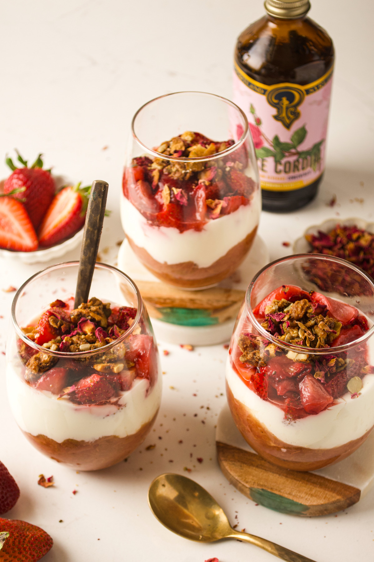 Three glasses of layered dessert with granola, yogurt, and strawberries on a white surface, surrounded by fresh strawberries, a spoon, and a bottle of rose cordial in the background.