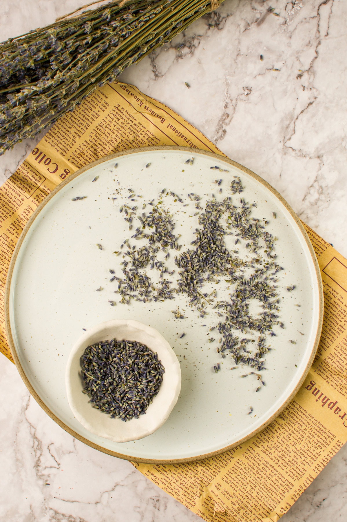 A white plate with scattered dried lavender buds and a small bowl filled with more lavender, placed on a yellow newspaper on a marble surface. A bundle of dried lavender rests in the corner.