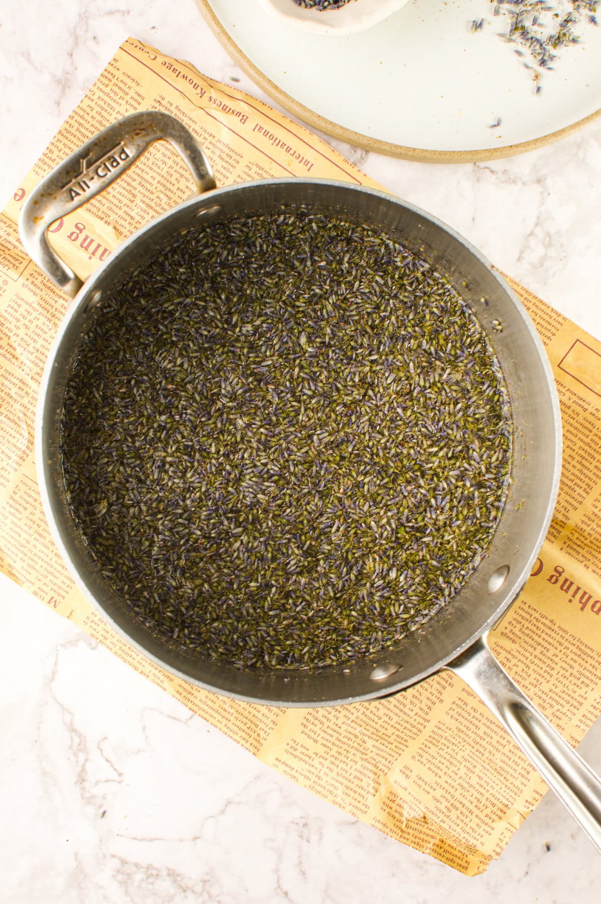 A stainless steel saucepan filled with a mixture of dried lavender buds and liquid sits on a newspaper on a marble surface.