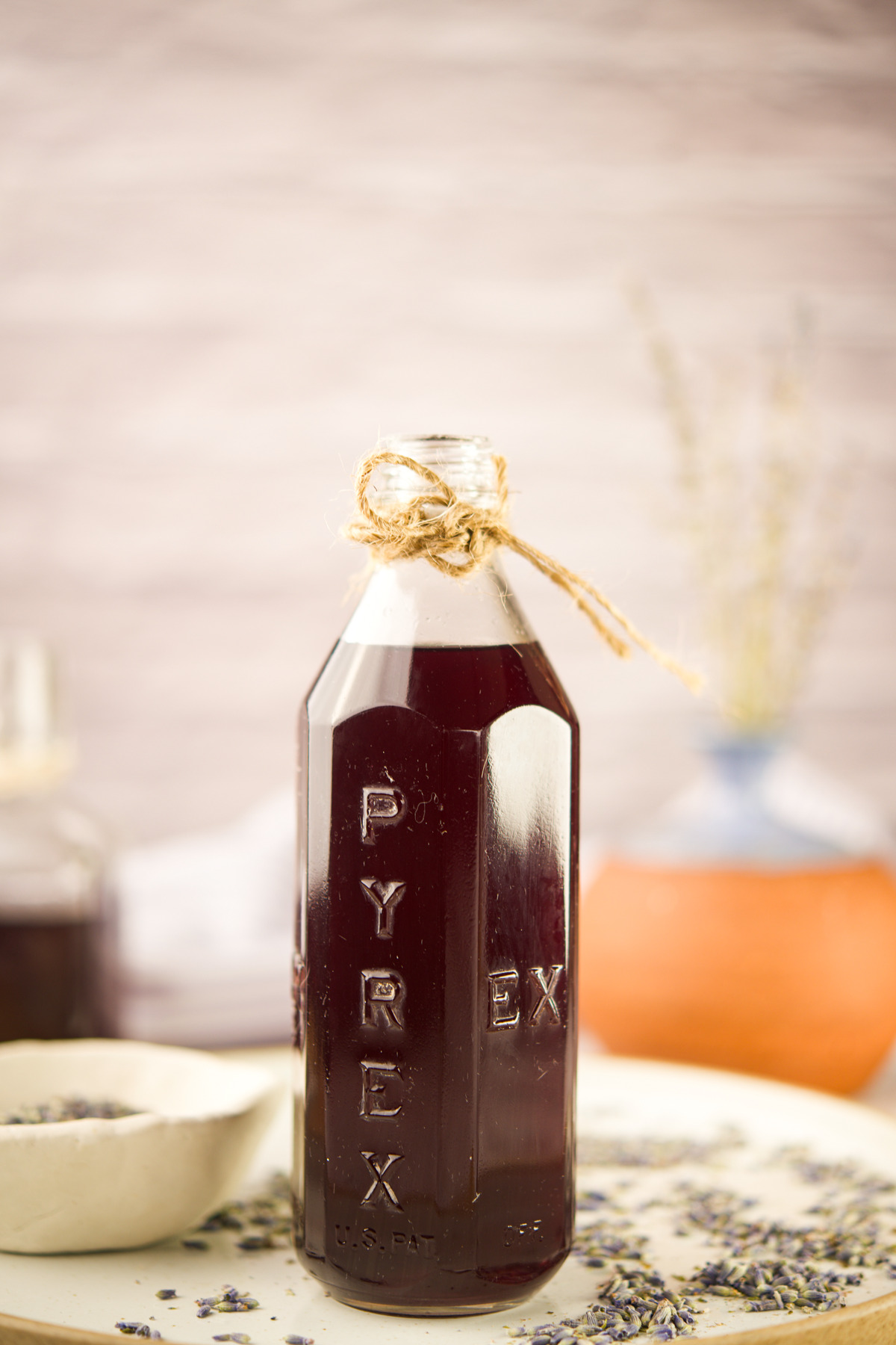 A glass bottle with Homemade Lavender Simple Syrup tied with twine at the neck, sits on a light surface with dried lavender scattered nearby; a blurred vase and objects are in the background.